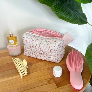 May include: A white quilted makeup bag with pink floral print and a pink and white gingham lining. The bag is open and has a pink makeup wipe inside. The bag is on a wooden table with a pink hairbrush, a clear glass bottle with a dropper, a white jar, and a yellow hair clip.