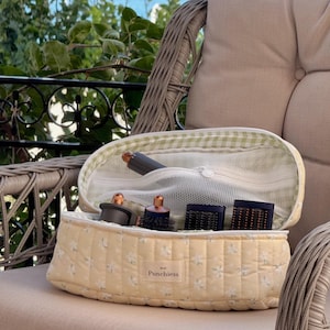 May include: A yellow floral quilted cosmetic bag with a green and white gingham interior, open to reveal hair styling tools. The bag is on a wicker chair with a neutral cushion, with a view of greenery and water in the background.