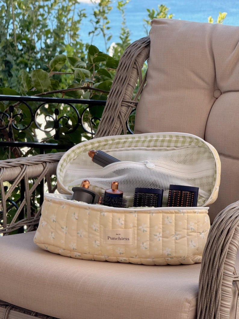 May include: A yellow floral quilted cosmetic bag with a green and white gingham interior, open to reveal hair styling tools. The bag is on a wicker chair with a neutral cushion, with a view of greenery and water in the background.