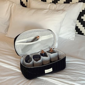 May include: Black oval cosmetic travel case with a striped interior and several compartments holding beauty tools. The case is open, revealing the contents. The case is on a white bed with white pillows and a black and white patterned headboard.