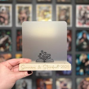 Puede incluir: Un expositor de acr&iacute;lico transparente con base de madera. El expositor presenta el texto "Sinners & Stardust 2025" y un dise&ntilde;o floral sobre un libro abierto. El fondo es una pared de im&aacute;genes enmarcadas.