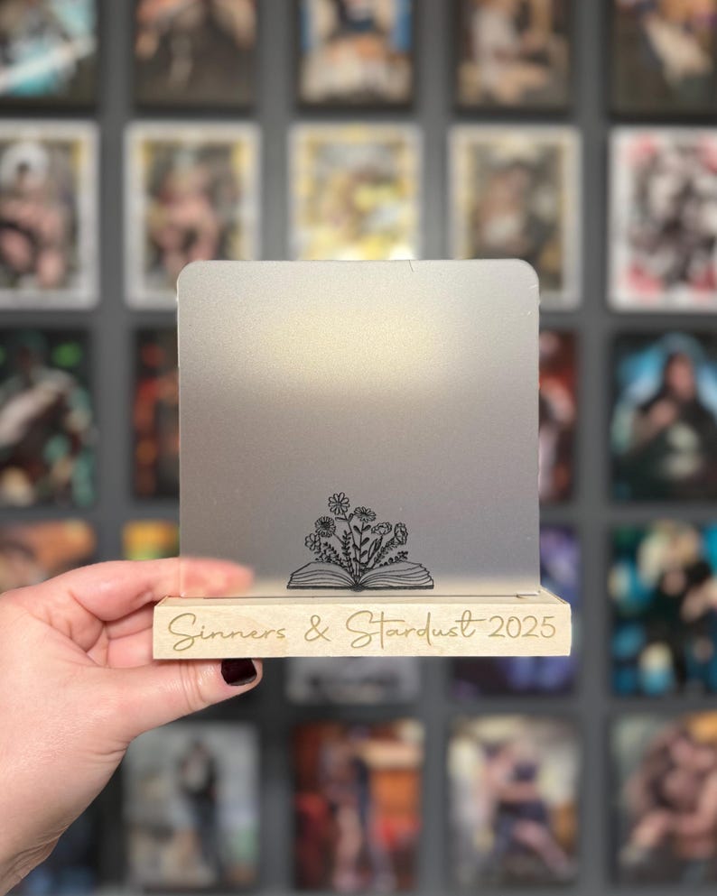 Puede incluir: Un expositor de acr&iacute;lico transparente con base de madera. El expositor presenta el texto "Sinners & Stardust 2025" y un dise&ntilde;o floral sobre un libro abierto. El fondo es una pared de im&aacute;genes enmarcadas.