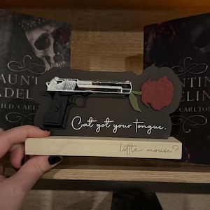 May include: A black wooden sign with a white silhouette of a gun and a red rose. The sign reads "Cat got your tongue. little mouse?"