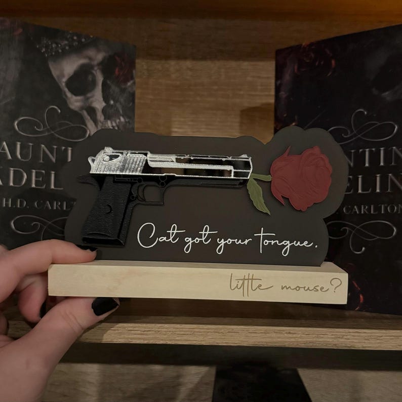 May include: A black wooden sign with a white silhouette of a gun and a red rose. The sign reads "Cat got your tongue. little mouse?"
