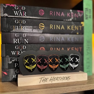 May include: A set of five black masks with neon green, yellow, orange, pink, and blue accents. The masks have a stitched smile and two X's for eyes. The masks are displayed on a black stand with the words "The Heathens" written on a wooden base. The masks are in front of a stack of books with the title "God of War" and the author "Rina Kent".