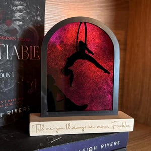 May include: A black silhouette of a person hanging from a hoop against a pink and purple glitter background. The silhouette is framed in a black arch and sits on a wooden base with the text "Tell me you'll always be mine, Freckles."