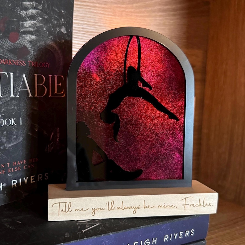 May include: A black silhouette of a person hanging from a hoop against a pink and purple glitter background. The silhouette is framed in a black arch and sits on a wooden base with the text "Tell me you'll always be mine, Freckles."