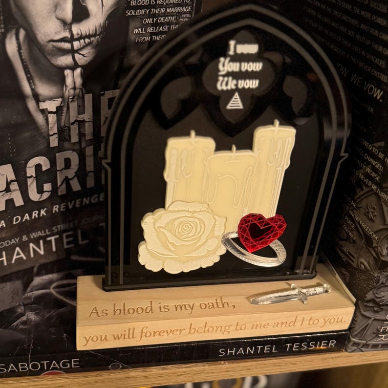 May include: A black and beige decorative piece with a gothic arch design. It features three cream-colored candles, a white rose, and a red heart-shaped gem ring. The base has engraved text and a small dagger. The top reads "I vow, You vow, We vow."