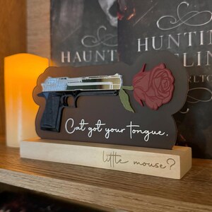 May include: A wooden sign with a black gun and a red rose, with the text "Cat got your tongue. little mouse?"