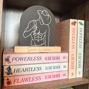 May include: A black and white line art illustration of a man with a hat, on a grey wooden sign with the text "Save a horse". The sign is propped up on a stack of four pink and white paperback books with the titles "Powerless", "Heartless", "Flawless", and "Reckless" by Elsie Silver.