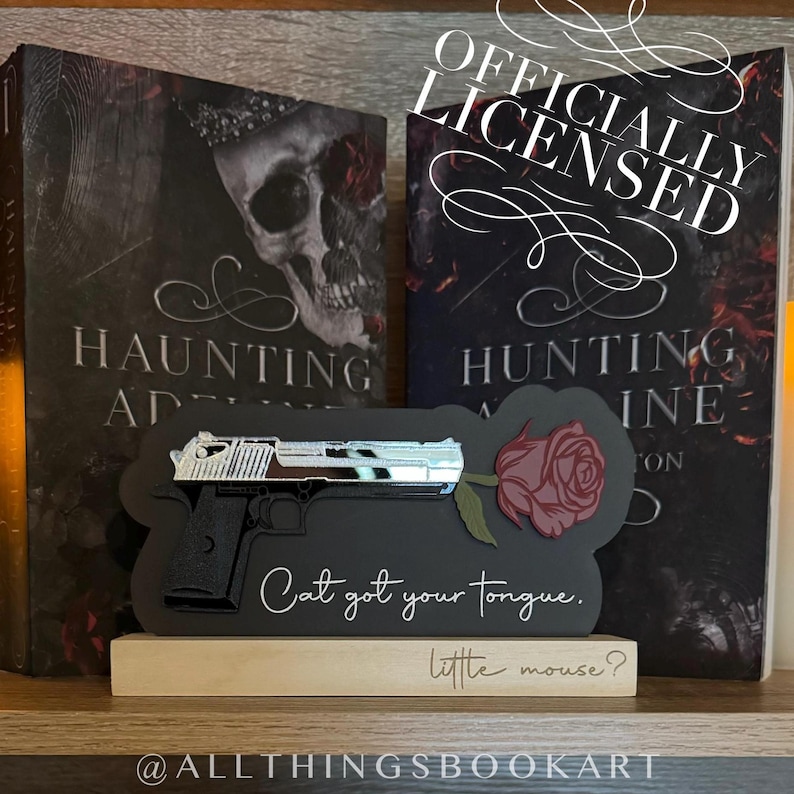 May include: A black and silver gun with a red rose attached to the barrel. The gun is on a black background with the text "Cat got your tongue. little mouse?"  The background is a bookshelf with two books titled "Haunting" and "Hunting".