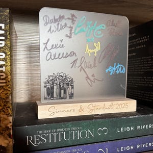 Puede incluir: Un bloque acr&iacute;lico transparente con firmas coloridas y un dise&ntilde;o floral de libros, sobre una base de madera. La base dice "Sinners & Stardust 2025". El bloque est&aacute; sobre una pila de libros.