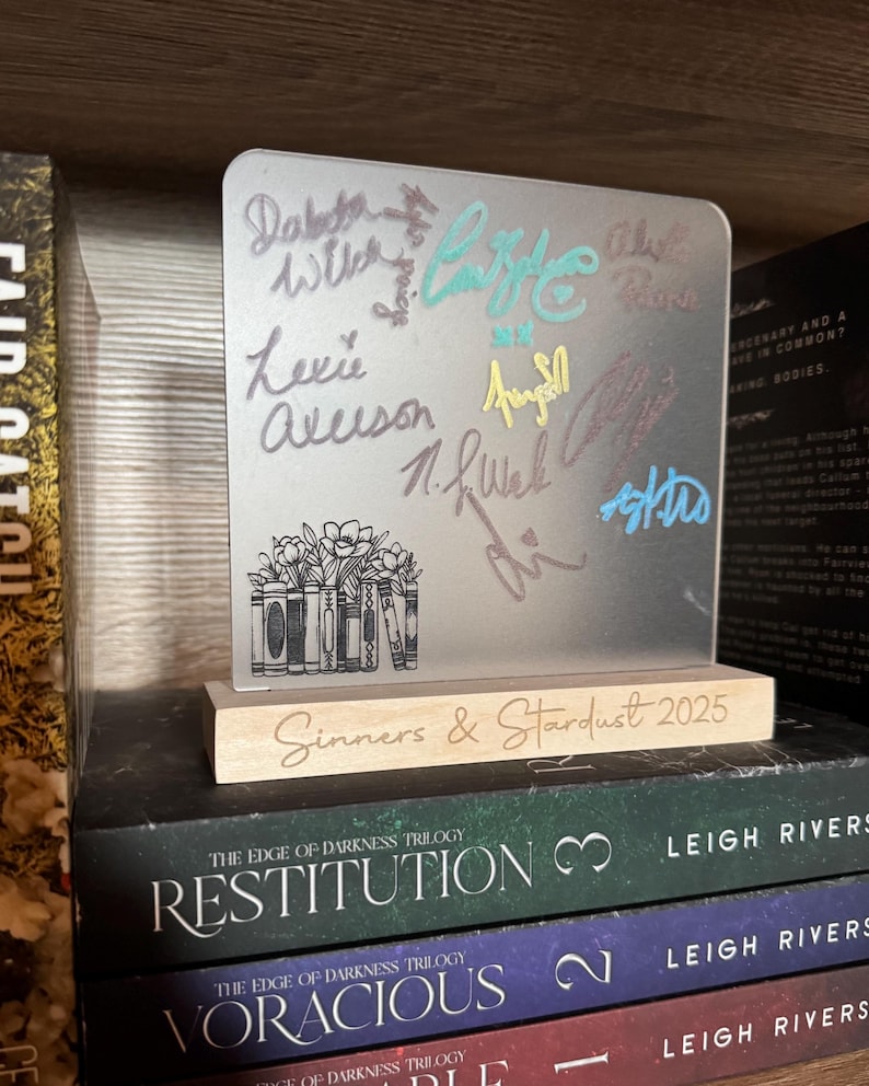 Puede incluir: Un bloque acr&iacute;lico transparente con firmas coloridas y un dise&ntilde;o floral de libros, sobre una base de madera. La base dice "Sinners & Stardust 2025". El bloque est&aacute; sobre una pila de libros.