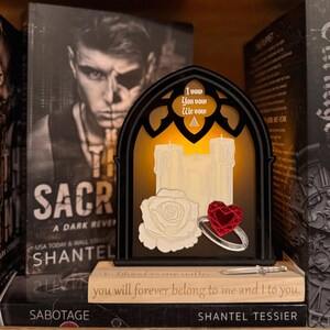 May include: A gothic-style decorative light with three faux candles, a white rose, and a ring with a red heart-shaped gem. The light has the text "I vow, You vow, We vow". The light sits on a stack of books.