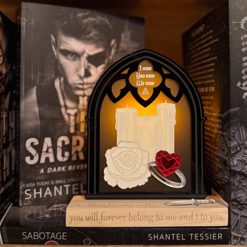 May include: A gothic-style decorative light with three faux candles, a white rose, and a ring with a red heart-shaped gem. The light has the text "I vow, You vow, We vow". The light sits on a stack of books.