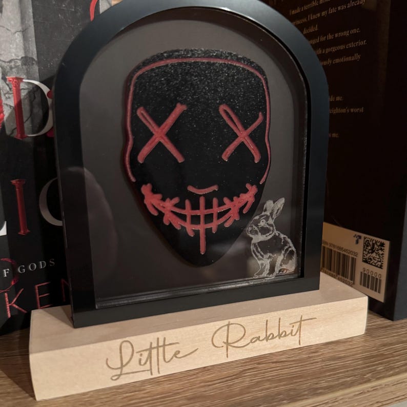 May include: A black and red mask with a stitched smile and X's for eyes is displayed in a black frame with a white rabbit figurine. The frame is sitting on a wooden base with the words "Little Rabbit" engraved on it.