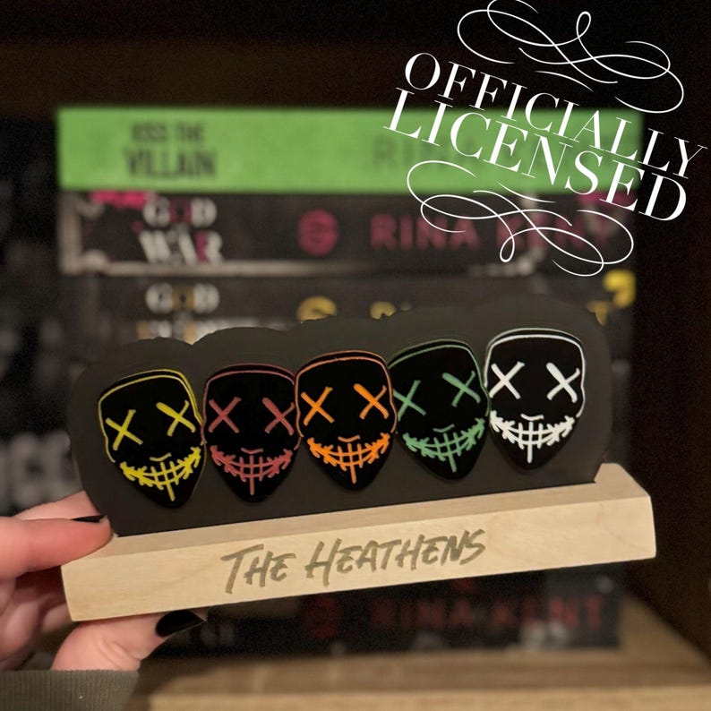 May include: A set of four black masks with neon green, yellow, orange, and red accents. Each mask has two black X's for eyes and a stitched mouth. The masks are displayed on a wooden stand with the text "The Heathens" written on it. The image also includes the text "Officially Licensed".