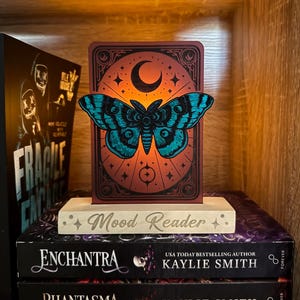 May include: A decorative illuminated card featuring a teal moth design, a crescent moon, and the words "Mood Reader." The card sits on a wooden base, placed on top of two books. The books are titled "Enchantra" and "Phantasma" by Kaylie Smith.