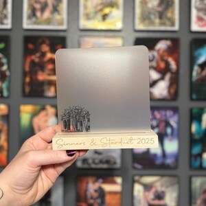 Puede incluir: Un soporte plateado para port&aacute;til con base de madera. La base tiene grabado el texto "Sinners & Stardust 2025". El soporte presenta un dise&ntilde;o floral y de libros. El soporte se sostiene frente a una pared de arte enmarcado.