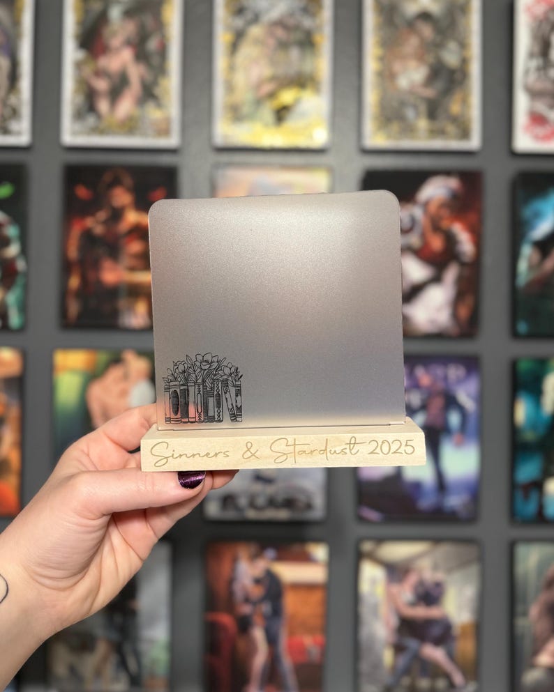 Puede incluir: Un soporte plateado para port&aacute;til con base de madera. La base tiene grabado el texto "Sinners & Stardust 2025". El soporte presenta un dise&ntilde;o floral y de libros. El soporte se sostiene frente a una pared de arte enmarcado.