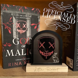 May include: A black and red mask with a stitched smile and X's for eyes is displayed in a black frame on a wooden base. The base is engraved with the words "Little Rabbit". The mask is from the book "God of Malice" by Rina Kent.
