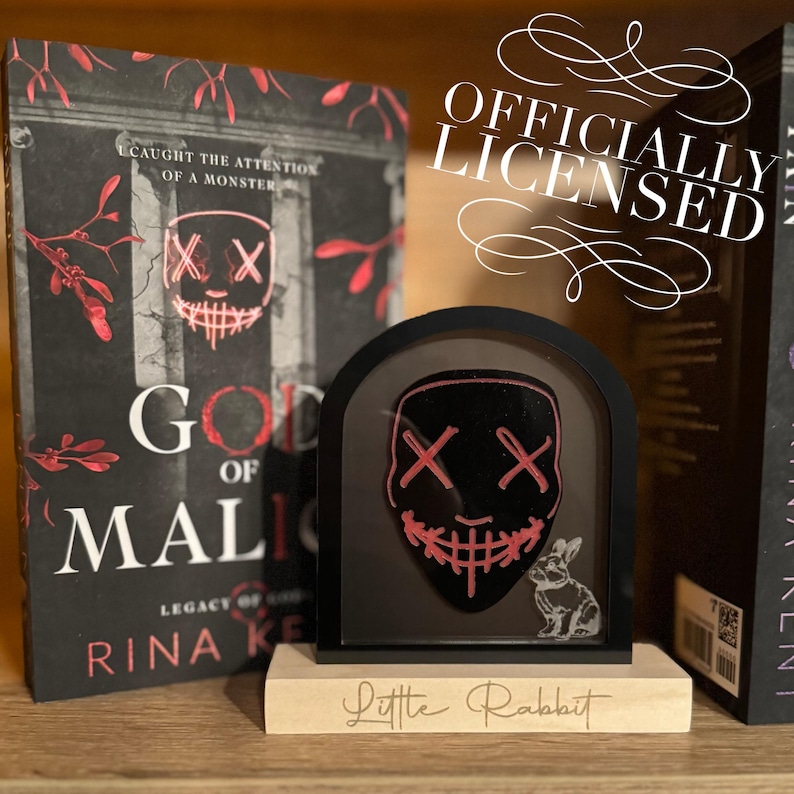 May include: A black and red mask with a stitched smile and X's for eyes is displayed in a black frame on a wooden base. The base is engraved with the words "Little Rabbit". The mask is from the book "God of Malice" by Rina Kent.