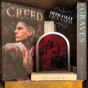 May include: A bookend with a red and black silhouette of a musician and drum kit, with the text "We need us." The bookend is placed between two books, one titled "CREED" and the other "GRAVES". The image also includes the text "OFFICIALLY LICENSED".