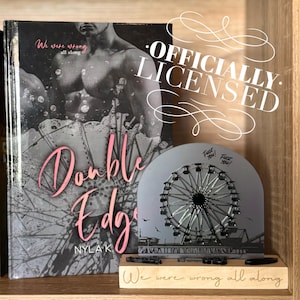 May include: A book titled "Double Edge" by Nyla K. is displayed next to a Ferris wheel-shaped decorative item. The book cover features a grayscale image of a person and a Ferris wheel. The decorative item has the words "Fuck You!" and "We were wrong all along" on it.