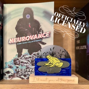 May include: A book titled "NEUROVANCE" with a dark figure on the cover, and a decorative frog pin on a wooden base that reads "I'll meet you at the pond." The pin features a yellow frog on lily pads.