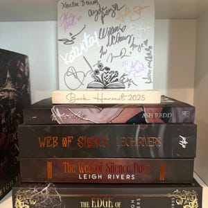 Puede incluir: Una pila de libros con cubiertas oscuras y letras doradas. Los t&iacute;tulos incluyen "Ash Redd", "Web of Silence" y "The Edge of Darkness". Un letrero blanco con firmas y el texto "Book Harvest 2025" se encuentra encima.