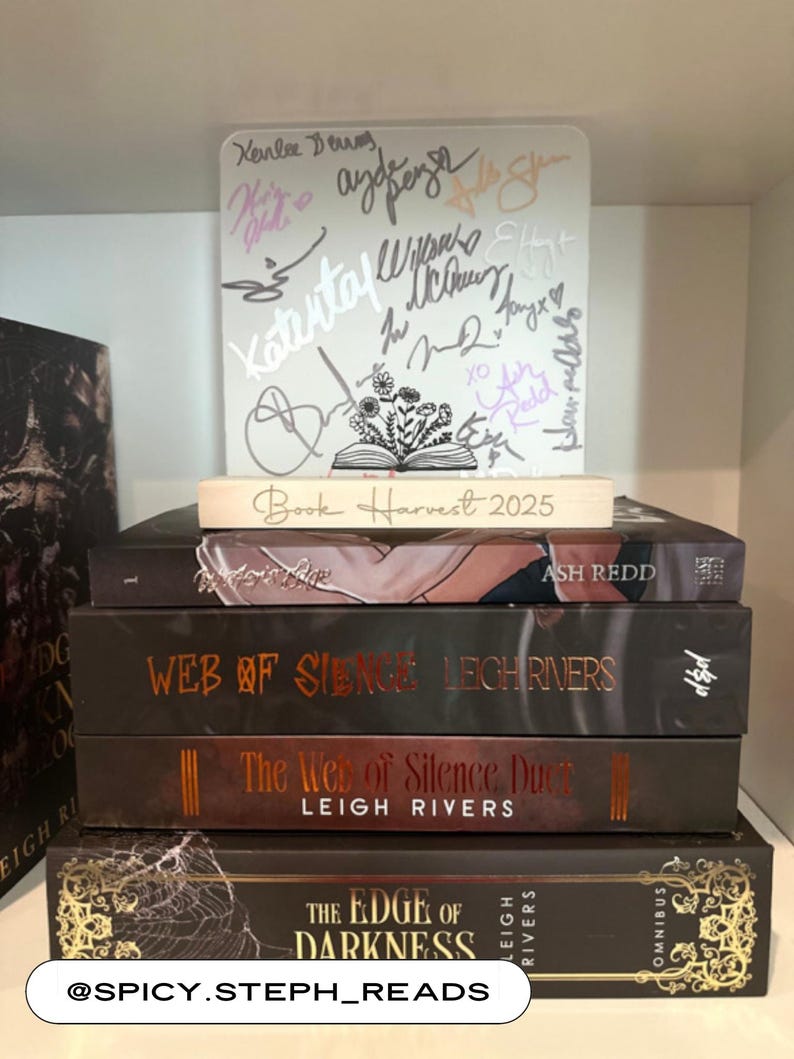 Puede incluir: Una pila de libros con cubiertas oscuras y letras doradas. Los t&iacute;tulos incluyen "Ash Redd", "Web of Silence" y "The Edge of Darkness". Un letrero blanco con firmas y el texto "Book Harvest 2025" se encuentra encima.