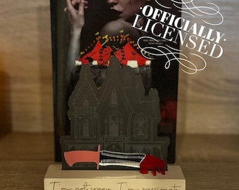 May include: A black and gray house-shaped bookend with a red and black knife sticking out of the side. The bookend is sitting on a wooden surface with the text "I'm not crazy. I'm passionate." and "@ALLTHINGSBBOOKART" written on it.