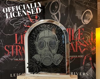 Puede incluir: Una caja de sombra enmarcada en metal plateado con un diseño de máscara de gas negra y gris. La caja de sombra está sentada sobre una base de madera con el texto "Run, little stranger." La caja de sombra está delante de dos libros, uno con el título "Little Stranger" y el otro con el título "Silence Duet".