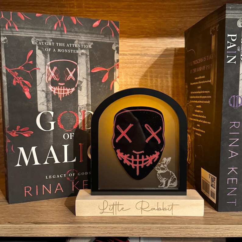 May include: A black and red mask with a smiling mouth and two X's for eyes is illuminated in a black and gold frame. The frame is on a wooden base with the words "Little Rabbit" written on it. The frame is in front of two books, one titled "God of Malice" and the other titled "Pain".