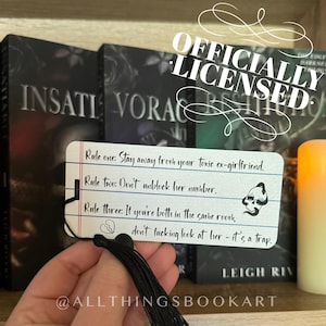 May include: A white bookmark with black text that reads "Rule one: Stay away from your toxic ex-girlfriend." A black tassel hangs from the bottom. The background includes book covers and a candle.