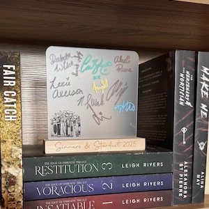 Puede incluir: Una exhibici&oacute;n de libros firmados y una placa conmemorativa. La placa, con firmas y el texto "Sinners & Stardust 2025", est&aacute; sobre una base de madera. Debajo, tres libros de "The Edge of Darkness Trilogy".