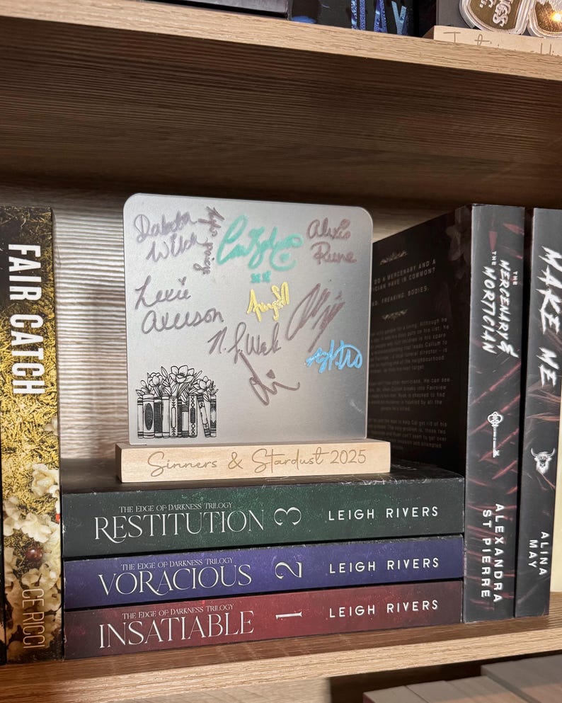 Puede incluir: Una exhibici&oacute;n de libros firmados y una placa conmemorativa. La placa, con firmas y el texto "Sinners & Stardust 2025", est&aacute; sobre una base de madera. Debajo, tres libros de "The Edge of Darkness Trilogy".