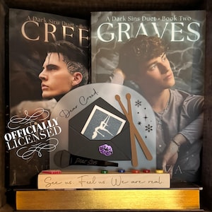 May include: A display featuring two books, "Cree" and "Graves," from "A Dark Sins Duet." A decorative piece includes an envelope with a photo, drumsticks, and a microphone. The text "Dear Creed" and "See us. Feel us. We are real" are visible.