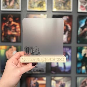 Puede incluir: Soporte de exhibici&oacute;n rectangular plateado con base de madera. La base tiene grabado el texto "Sinners & Stardust 2025". El soporte presenta un dise&ntilde;o grabado de libros y una calavera.