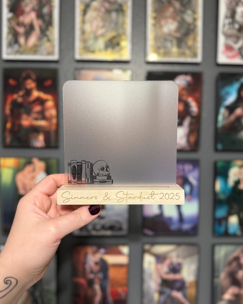 Puede incluir: Soporte de exhibici&oacute;n rectangular plateado con base de madera. La base tiene grabado el texto "Sinners & Stardust 2025". El soporte presenta un dise&ntilde;o grabado de libros y una calavera.