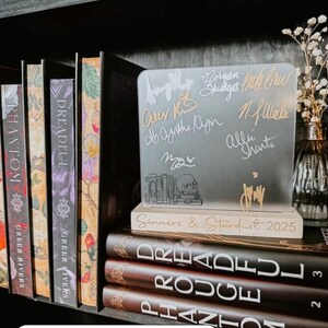 Puede incluir: Libros en una estanter&iacute;a, incluyendo "Phantom" y "Dreadful". Un letrero de acr&iacute;lico transparente con firmas y el texto "Sinners & Stardust 2025" se encuentra encima de los libros. Un jarr&oacute;n con flores secas es visible.
