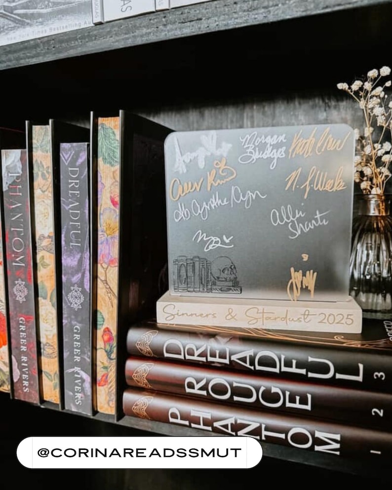 Puede incluir: Libros en una estanter&iacute;a, incluyendo "Phantom" y "Dreadful". Un letrero de acr&iacute;lico transparente con firmas y el texto "Sinners & Stardust 2025" se encuentra encima de los libros. Un jarr&oacute;n con flores secas es visible.