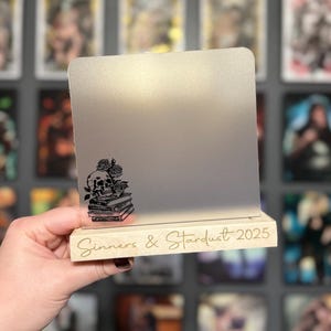 Puede incluir: Un letrero decorativo con una superficie gris claro y una base de madera. El letrero presenta un dise&ntilde;o de calavera y rosas negras sobre una pila de libros. Las palabras "Sinners & Stardust 2025" est&aacute;n grabadas en la base.