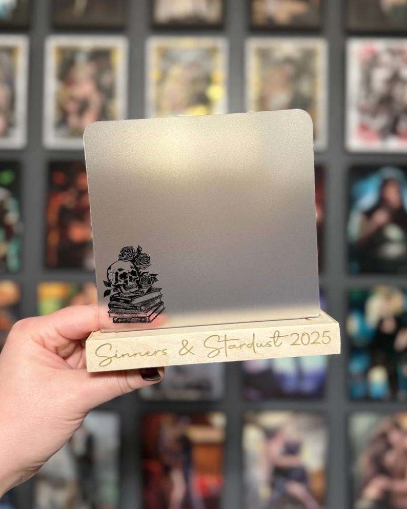 Puede incluir: Un letrero decorativo con una superficie gris claro y una base de madera. El letrero presenta un dise&ntilde;o de calavera y rosas negras sobre una pila de libros. Las palabras "Sinners & Stardust 2025" est&aacute;n grabadas en la base.
