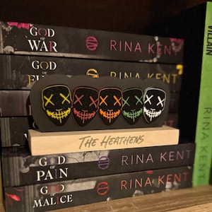 May include: A stack of books with the title "God of War", "God of Fire", "God of Pain", and "God of Malice" by Rina Kent. A wooden stand with the text "The Heathens" holds five colorful masks with the letter "X" on them.