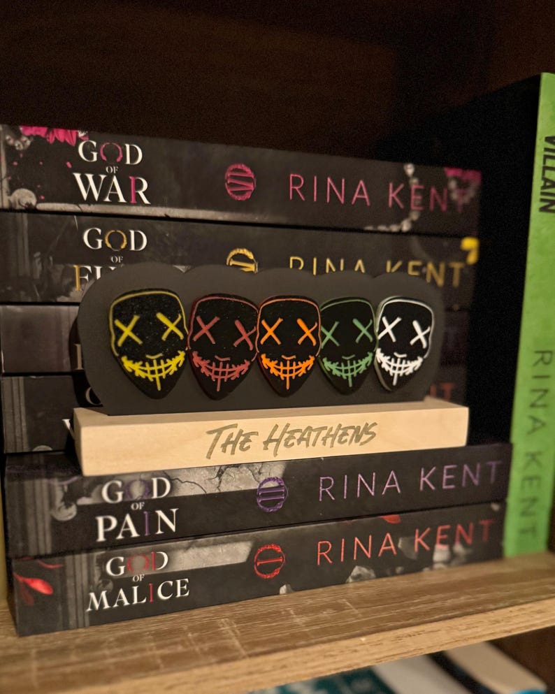 May include: A stack of books with the title "God of War", "God of Fire", "God of Pain", and "God of Malice" by Rina Kent. A wooden stand with the text "The Heathens" holds five colorful masks with the letter "X" on them.