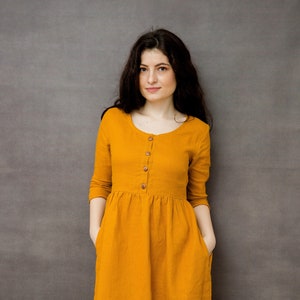 May include: A mustard yellow linen dress with a button-up front and 3/4 sleeves. The dress has a gathered waist and pockets on the sides.