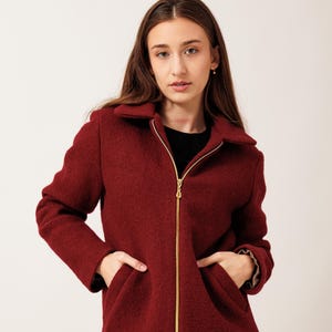 Op de afbeelding: Bordeaux wollen jasje met een gouden rits en twee voorzakken. De jas heeft een kraag en lange mouwen. Het model draagt een zwarte top en een zwarte jeans. Stijlvolle bovenkleding.