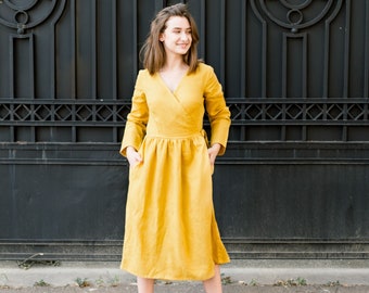 Pode incluir: Uma mulher vestindo um vestido amarelo de amarrar com mangas compridas e uma saia franzida. Ela está em pé em frente a um portão de metal preto e está olhando para a direita.