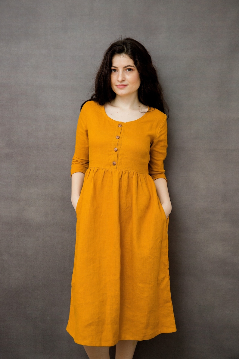 May include: A mustard yellow linen dress with a button-up front and 3/4 sleeves. The dress has a gathered waist and pockets on the sides.